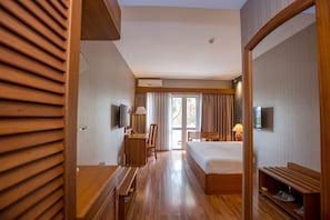 A hotel room with a wooden floor, a bed, a desk with a chair, a TV, and a window with curtains.