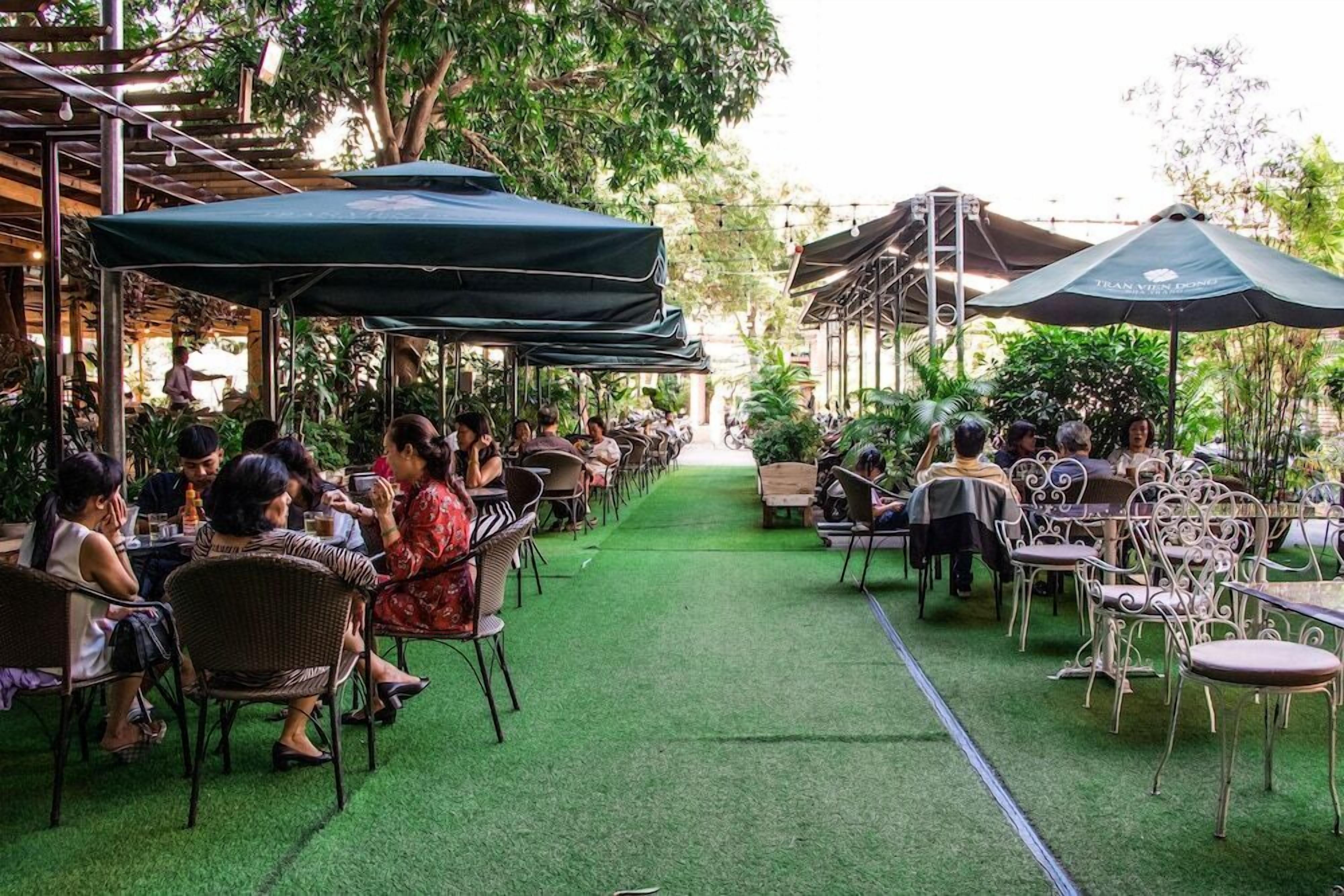An outdoor dining area with tables and chairs, covered by umbrellas, surrounded by greenery.
