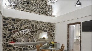 A dining area with a stone wall, a glass table, and a sofa with cushions. - Villa Murat (Santorini)
