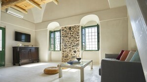A spacious living room with a high ceiling, a stone wall, and a television mounted on the wall. - Villa Murat (Santorini)