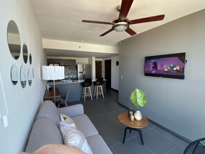 Ein modernes Wohnzimmer mit grauem Sofa, Deckenventilator, Fernseher und einer Küche im Hintergrund.
