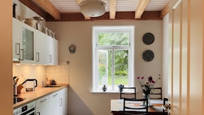 Eine Küche mit weißen Schränken, einer Holz Arbeitsplatte, einem Esstisch mit Stühlen, einem Fenster mit Blick ins Grüne und einer Wanduhr.