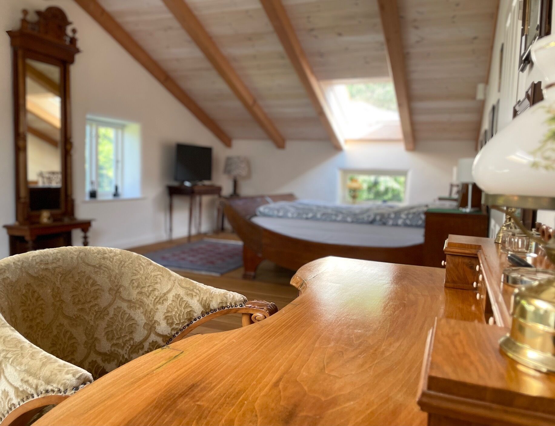 A room with a wooden table, a chair, a bed, a mirror, and a lamp.