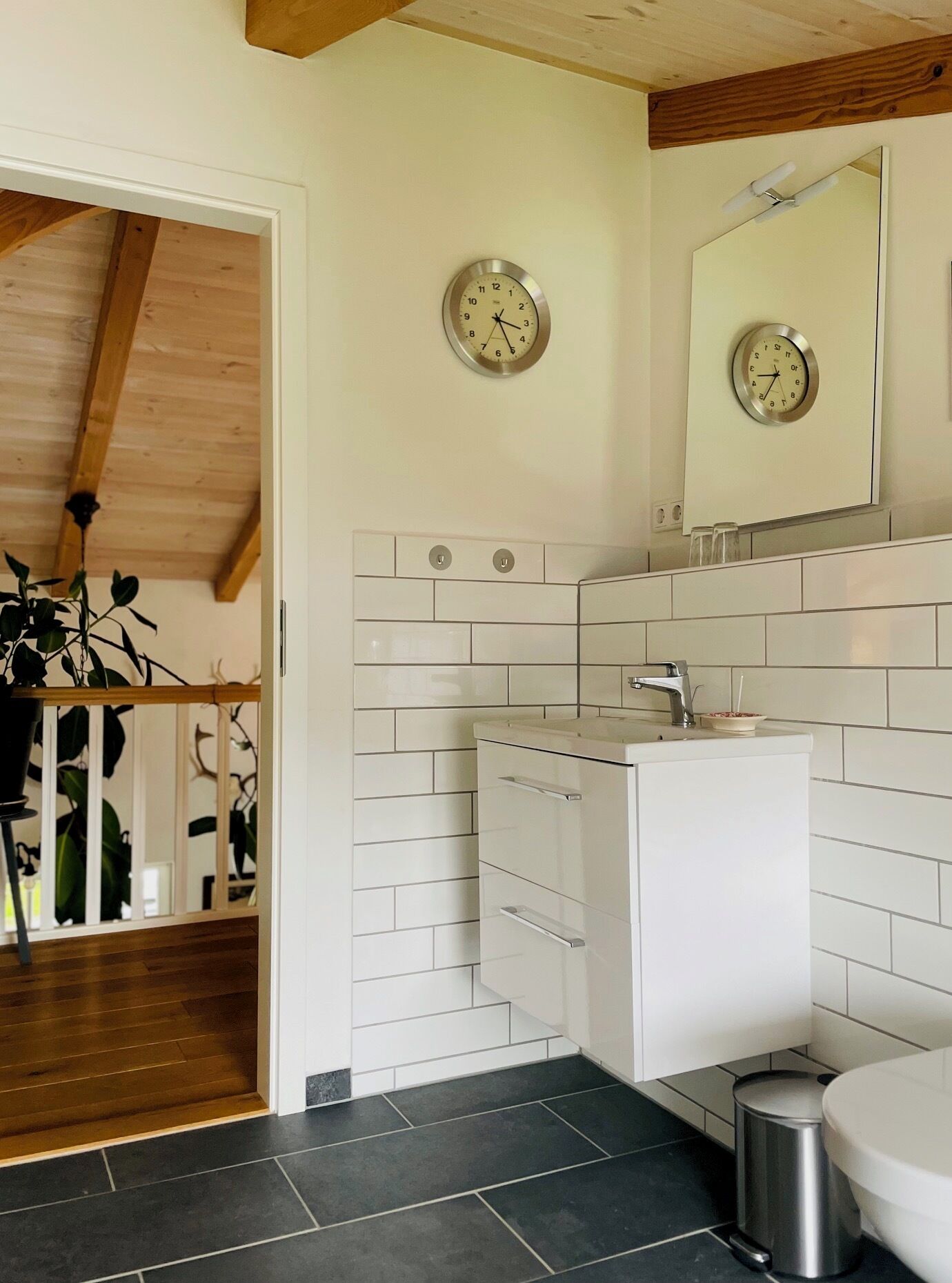 A bathroom with white tiles, a sink, a mirror, and two clocks on the wall.