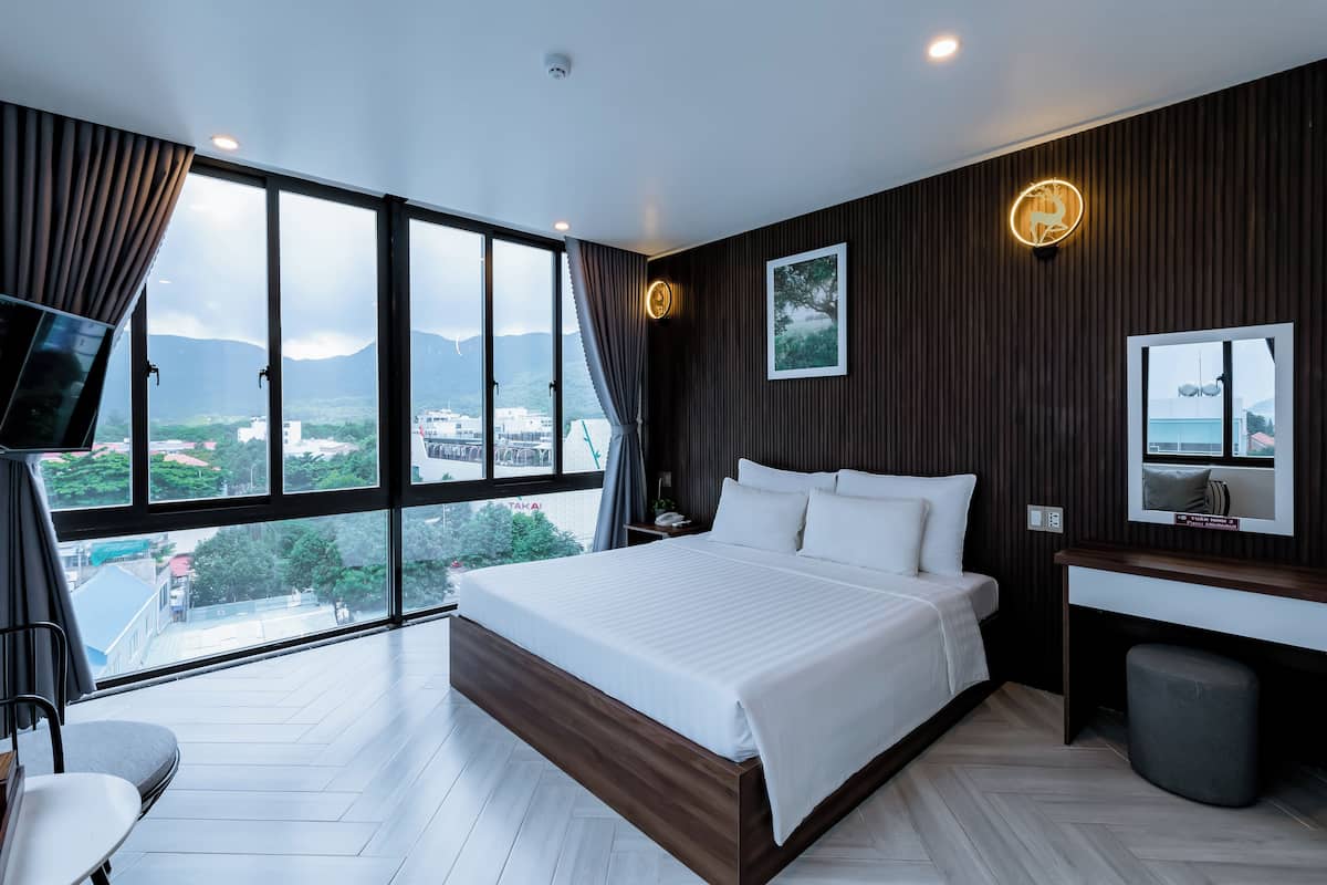 Una habitación de hotel moderna con una cama grande, un escritorio y vistas de la ciudad a través de amplias ventanas.
