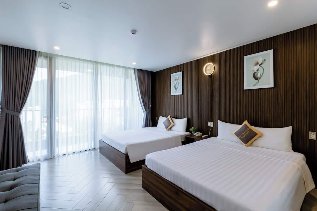 Habitación de hotel con dos camas, un ventanal grande con cortinas, un panel de pared de madera y una mesita de noche con lámpara.