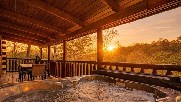 Une terrasse en bois avec un jacuzzi, des sièges d’extérieur et une vue sur les arbres et le coucher de soleil.