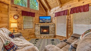 L’intérieur chaleureux d’un chalet avec une cheminée en pierre, des canapés en cuir, un téléviseur et une horloge au mur.