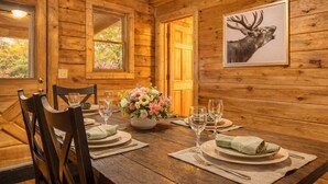 Une salle à manger rustique, aménagée dans une cabane en bois, avec une table dressée pour deux personnes, une tête de cerf empaillée accrochée au mur et une fenêtre donnant sur la forêt.