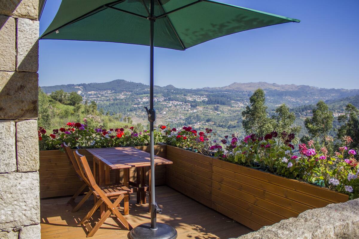 A patio with a wooden table and chairs, a green umbrella, and a flower bed with various flowers.