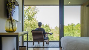 A modern bedroom with a large bed, a wooden chair, and a view of greenery through a large window.