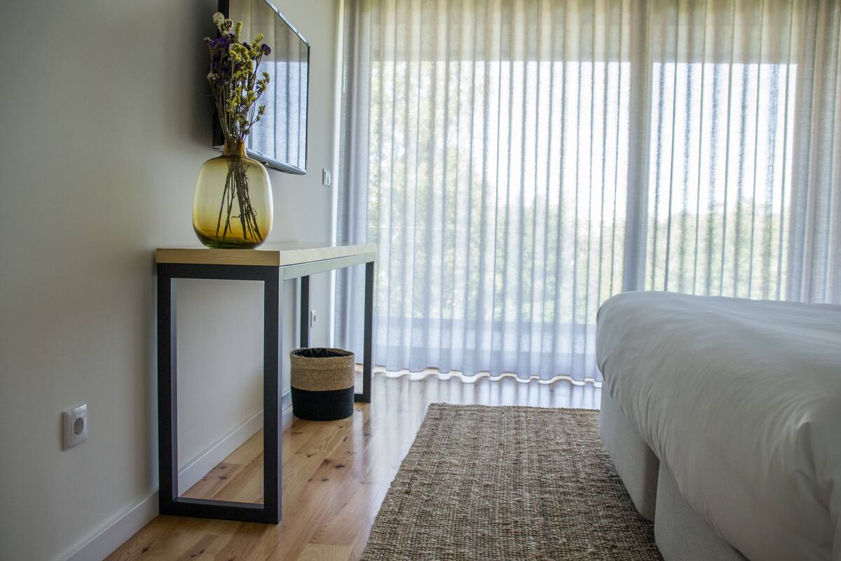A bedroom with a bed, a bedside table with a vase, a mirror, and a window with sheer curtains.