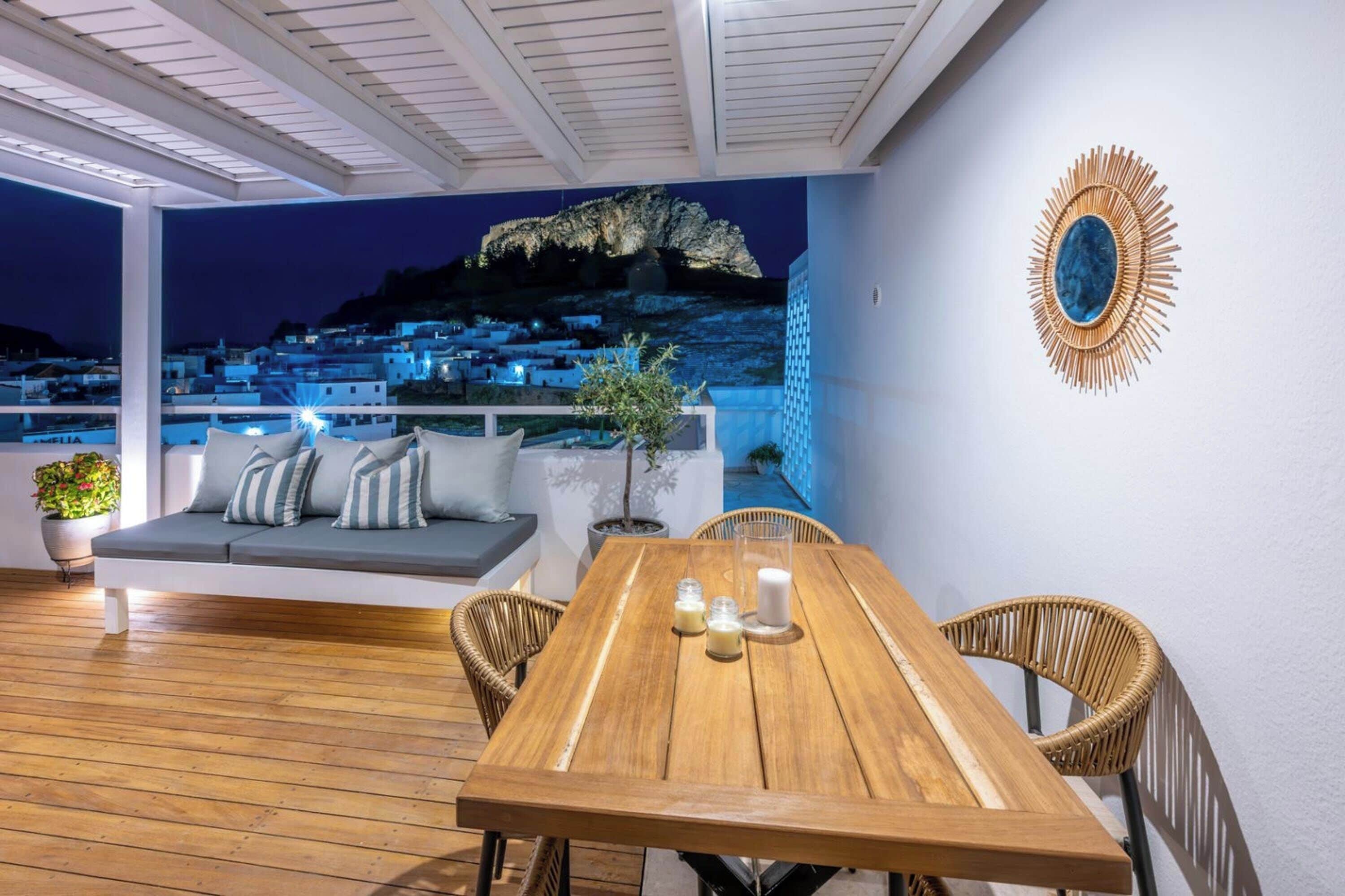 A wooden deck with a table and chairs, a sofa with cushions, a wall-mounted decorative mirror, and a view of a coastal village at night.