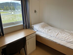 A small, single-bed room with a desk and chair, a window with curtains, and a view of a road and greenery.