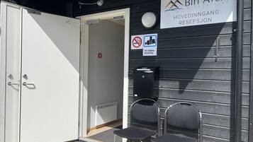 An entrance to a venue with a sign reading "Biri Arena Hovedindringang Resepsjon". There are two chairs outside, a door, and a no-smoking sign.