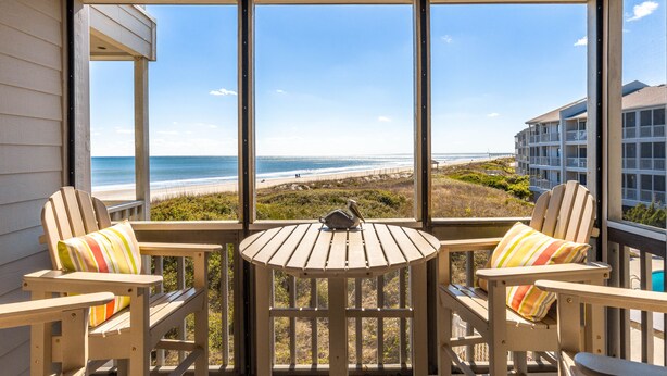 A balcony with two Adirondack chairs and a small table, overlooking a beach and ocean.