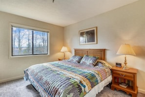 Quarto com uma cama grande, duas luminárias de cabeceira, mesa de cabeceira com livros, quadro na parede e janela com vista para árvores.