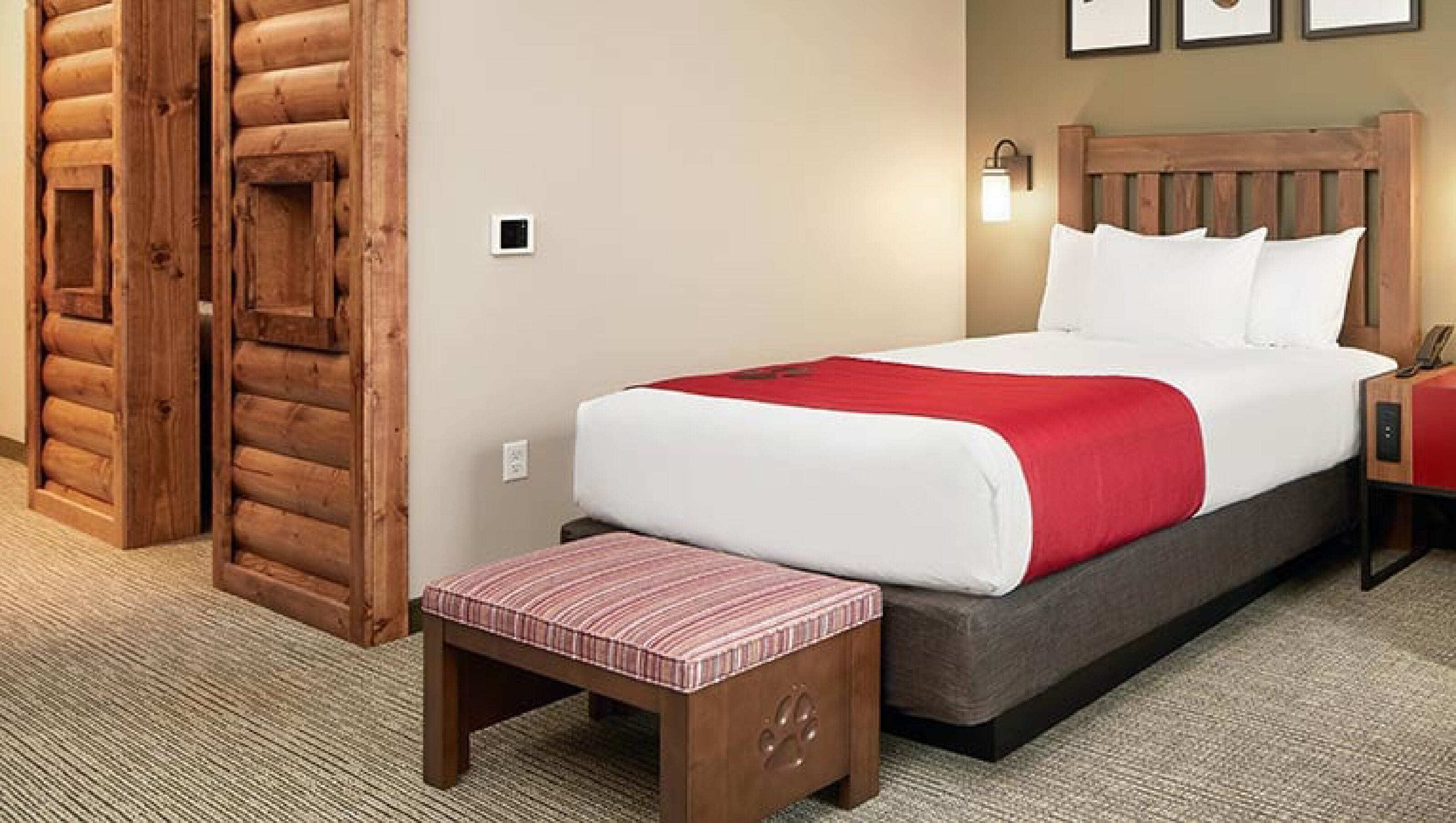 Habitación de hotel con pared de troncos de madera, cama con ropa de cama blanca y roja, un pequeño banco y una mesita de noche con una lámpara.