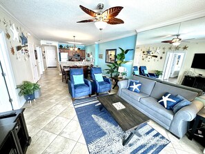 A spacious living room with a blue sofa, matching armchairs, and a coffee table. There is a kitchen in the background with a dining area. - Nice, Beautiful, Beach-Front, Condo with Large Balcony, Stay at Ocean Reef! (Panama City Beach)