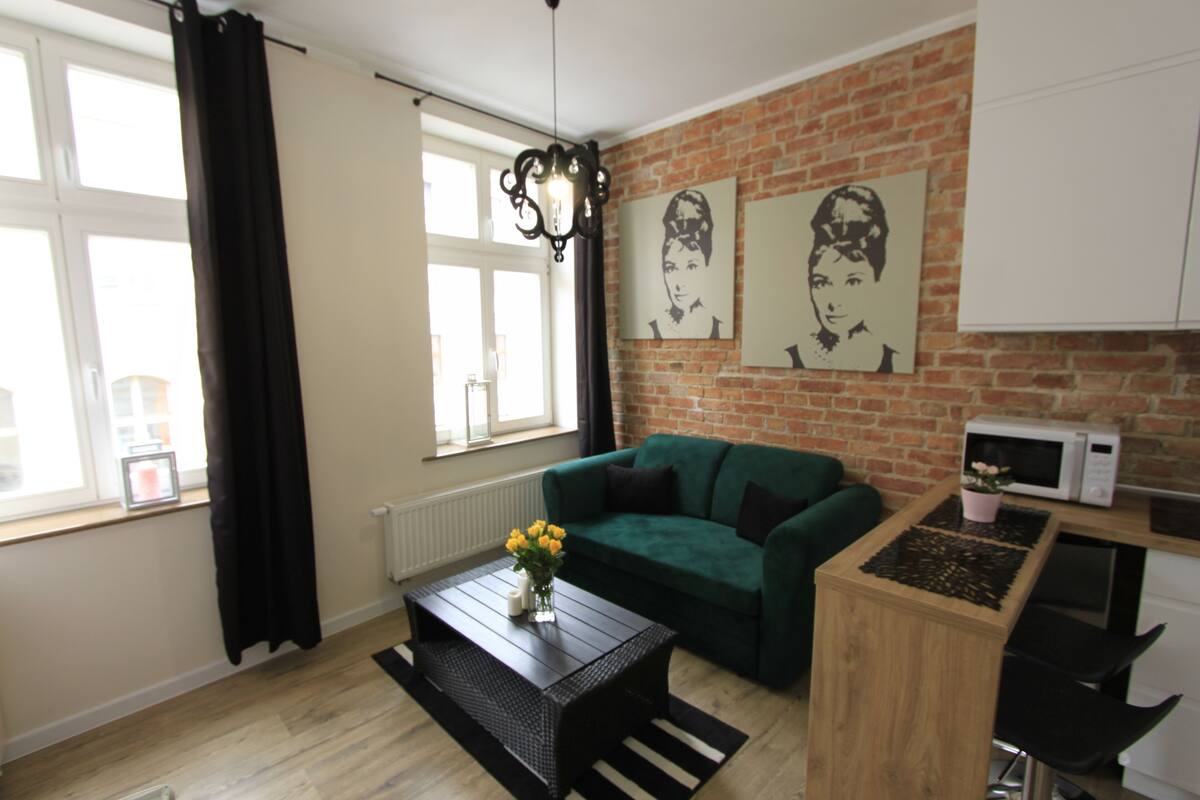 Sala de estar com sofá verde, mesa de centro com um vaso de flores amarelas, tapete listrado em preto e branco, parede de tijolos e janela com cortinas pretas.