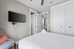 A bedroom with a bed, a TV mounted on the wall, a chair, and a desk with a computer.