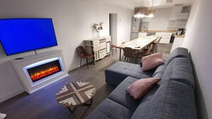 A modern living room with a grey sofa, a flat-screen TV, a fireplace, and a dining area in the background.