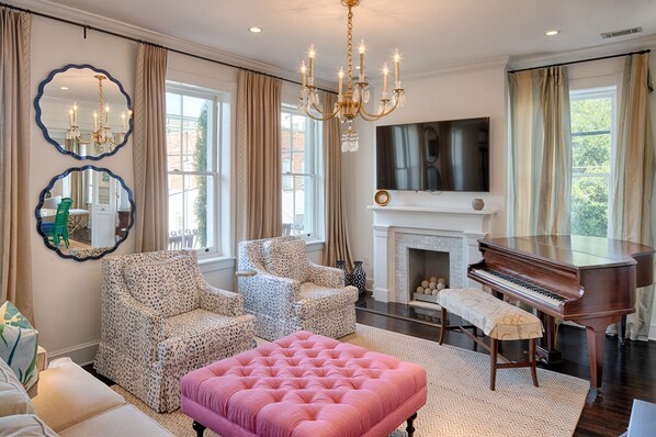 A living room with a piano, two patterned armchairs, a pink ottoman, a fireplace, and two wall-mounted mirrors.