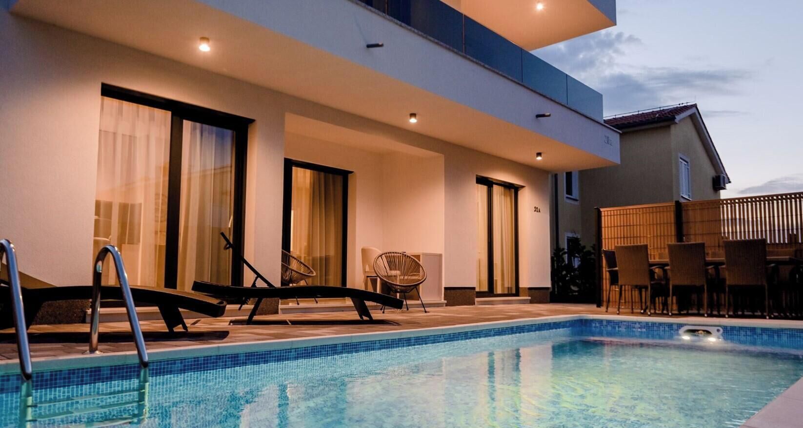 A modern outdoor pool area with lounge chairs and a dining table, surrounded by a contemporary building with large windows and doors.