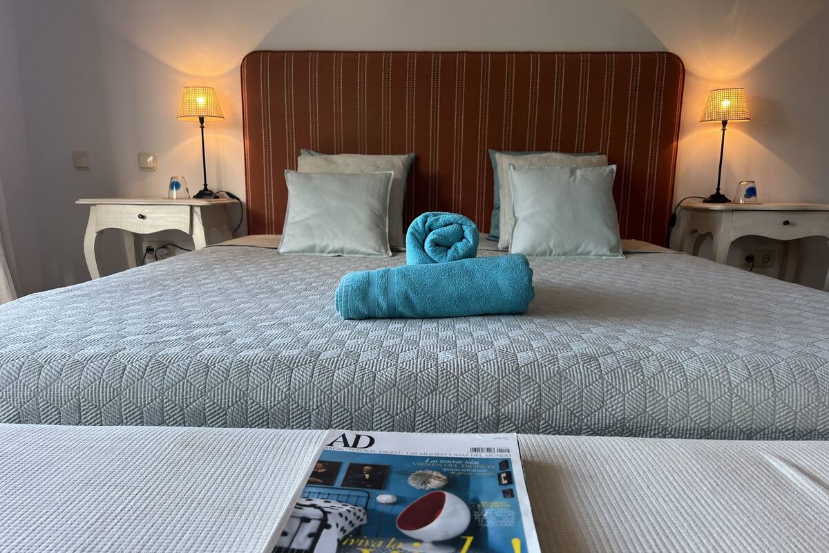 Un lit bien fait, avec une serviette pliée, deux lampes de chevet et un magazine posé sur le lit.