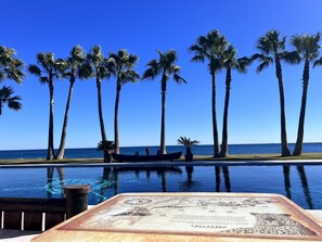 Una piscina con cubierta de madera, una placa conmemorativa, palmeras y vistas al océano.