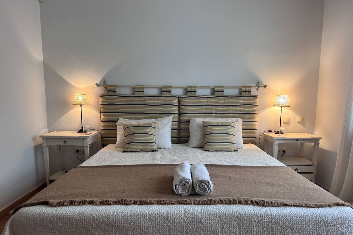 Un lit bien fait, avec une tête de lit à rayures, deux tables de chevet avec des lampes et une paire de serviettes enroulées posées sur le lit.
