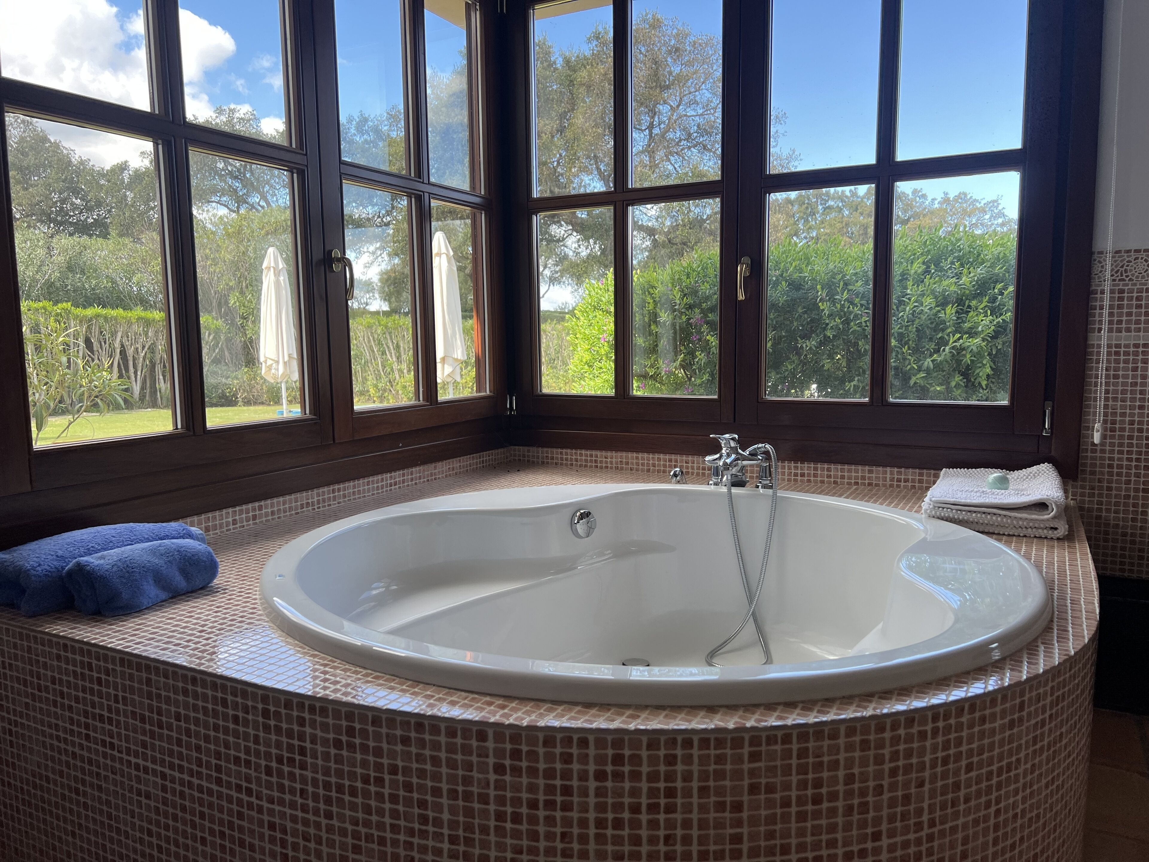 Un baño con una bañera grande, una ventana que da a zonas verdes y una toalla azul al lado.