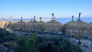A beach with thatched umbrellas and lounge chairs, a pier in the background, and a clear blue sky. - B&B Villa Maria Guest Rooms - San Roque Golf - Sotogrande (San Roque)