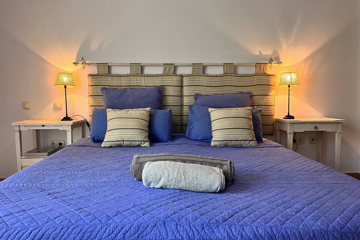 Un lit recouvert d’une couette bleue, deux tables de chevet avec des lampes et une tête de lit à rayures horizontales.