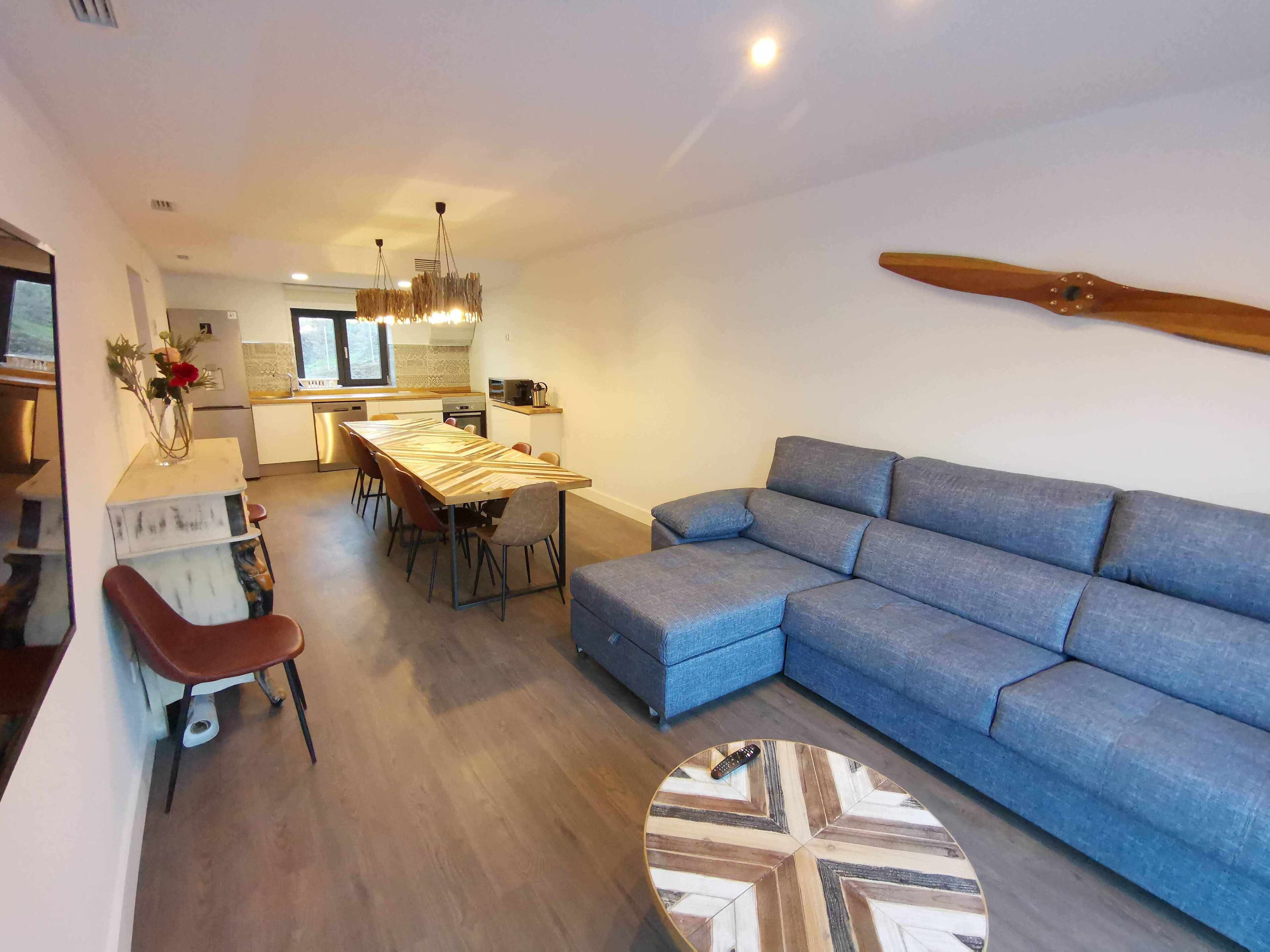 Ein modernes Wohnzimmer mit einem blauen Sofa, einem Holzesstisch und einer Küche im Hintergrund.