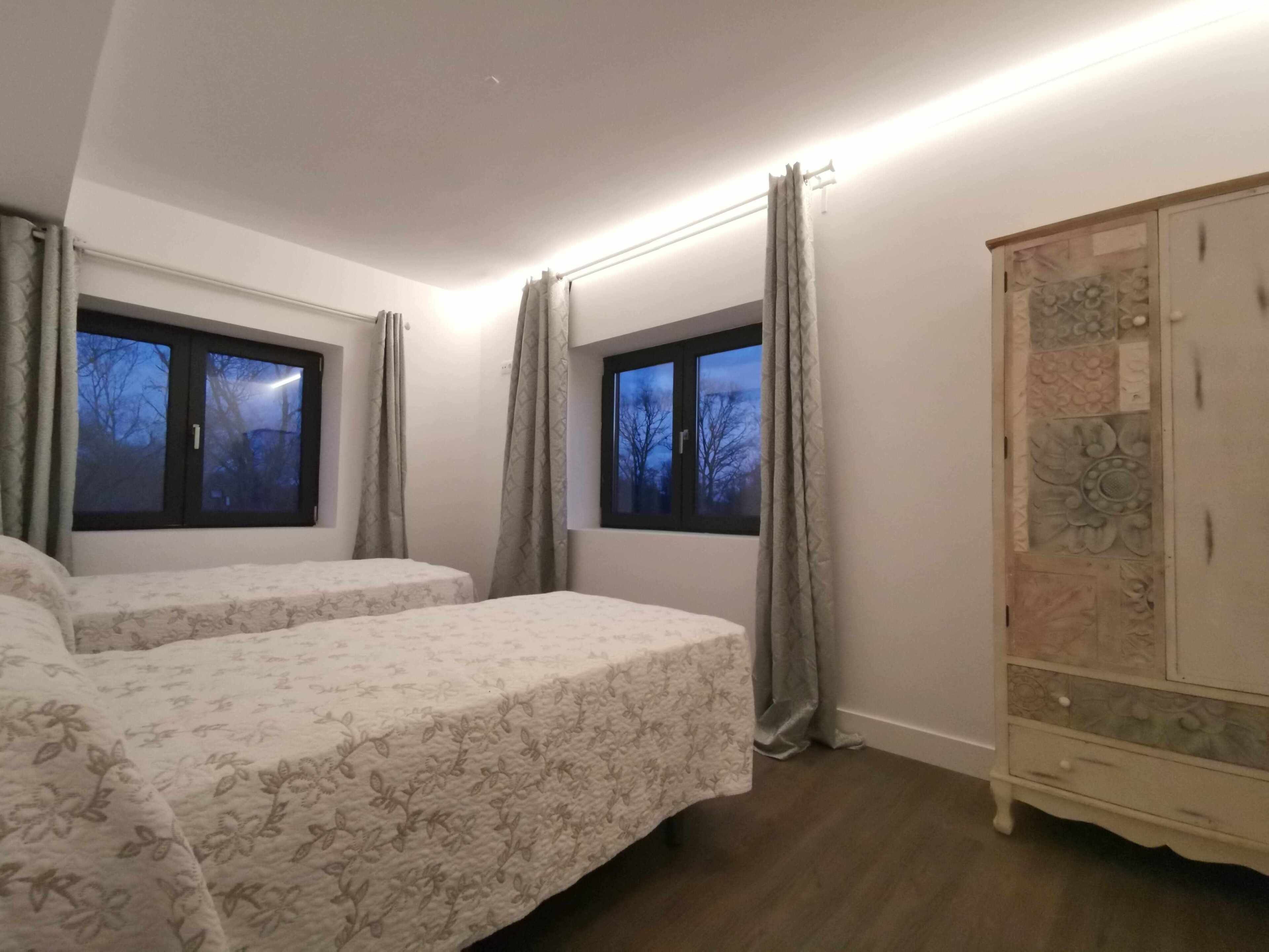 A bedroom with two beds, a wardrobe, and two windows with curtains.