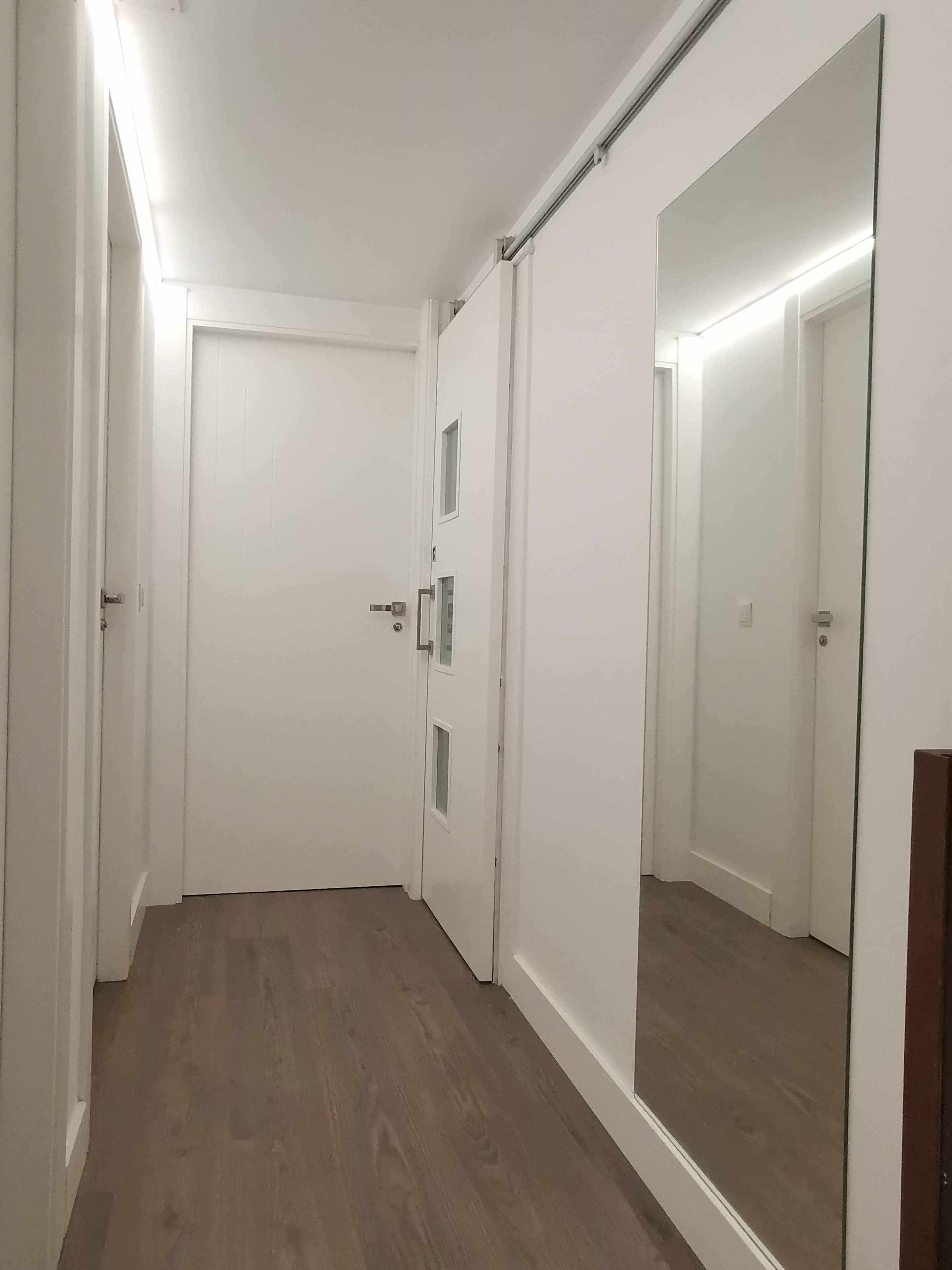 Un pasillo con piso de madera, paredes blancas y un espejo grande que refleja la puerta y una lámpara.