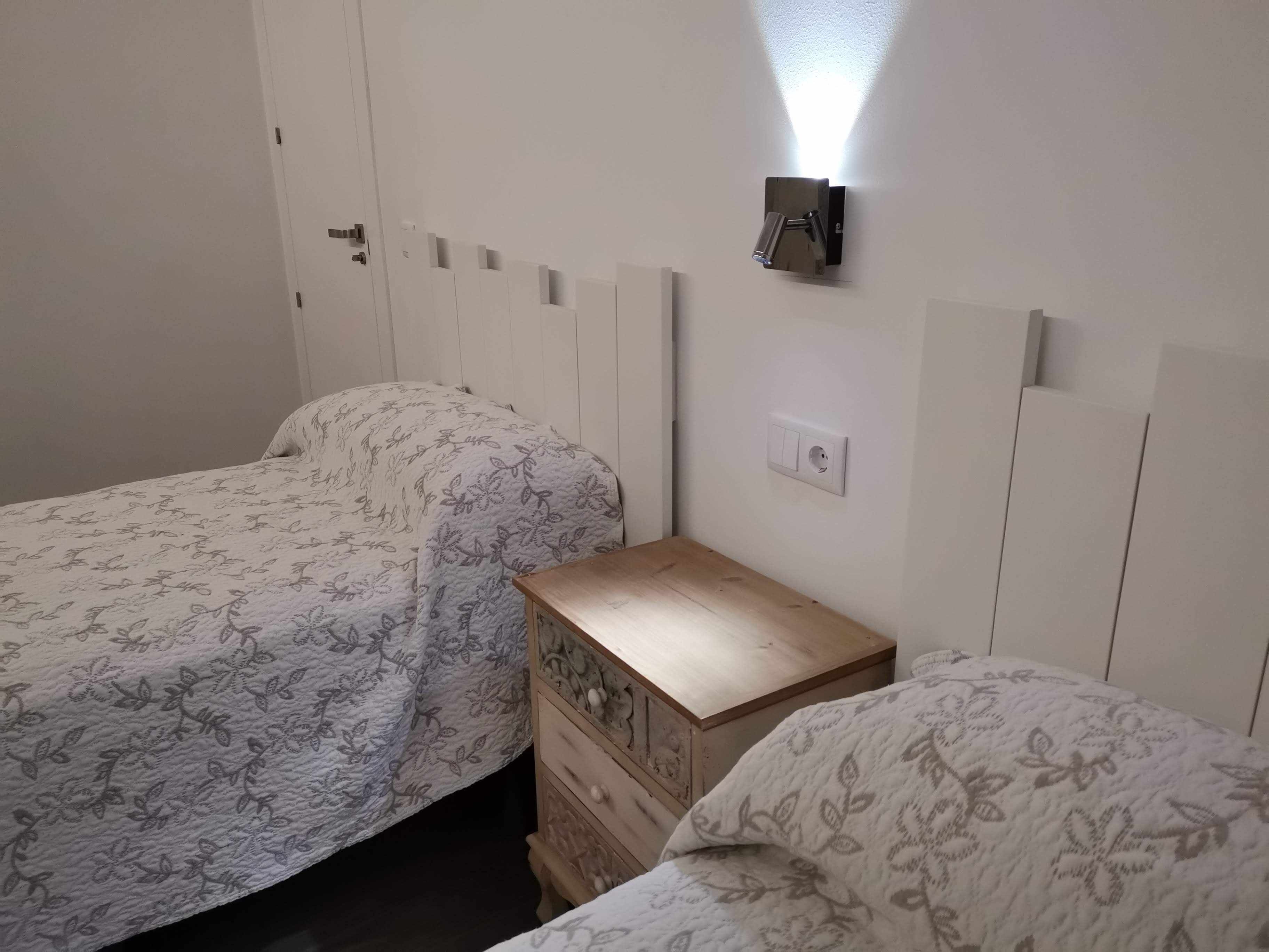 A bedroom with two single beds, a wooden nightstand, a wall-mounted light, and a power outlet.