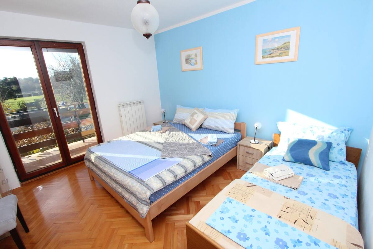 Ein Schlafzimmer mit einem hölzernen Bett, einem Nachttisch, einem Sessel und Blick durch eine Schiebetür.