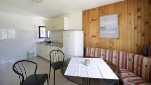 Una cocina compacta con armarios blancos, un refrigerador y un comedor con una mesa de vidrio y dos sillas. Se observa una pared de madera con un cuadro.