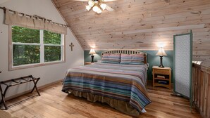 Un dormitorio con techo inclinado, una cama con ropa de cama a rayas, dos mesitas de noche con lámparas, una ventana con vistas a árboles y una cruz en la pared.