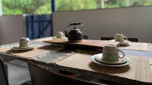 A table set with teapots and cups, a kettle, and a view of greenery through a window.