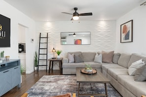 A modern living room with a sofa, coffee table, and a ceiling fan. There is a kitchen area in the background.