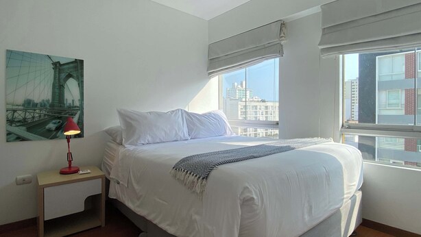 Une chambre à coucher avec un lit, une table de chevet, une lampe, une fenêtre donnant sur des immeubles et un tableau accroché au mur.