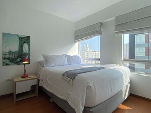Een slaapkamer met een bed, nachtkastje, lamp, raam met uitzicht op gebouwen en een schilderij aan de muur.