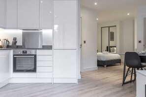 A modern kitchen with white cabinetry and a built-in oven, leading to a bedroom with a bed and a desk. - Solihull Studio • WiFi • NEC • BHX (Solihull)
