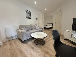 A modern, well-lit living room with a grey sofa, a round coffee table, and a black chair. A bed with a white headboard is visible in the background. - Solihull Studio • WiFi • NEC • BHX (Solihull)