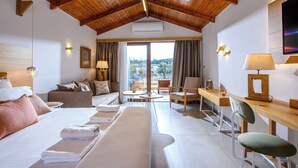 A hotel room with a wooden ceiling, a bed with white linens, a sofa, a desk with a chair, and a view of the outdoors through a large window. - Caldera Creta Paradise (Platanias)