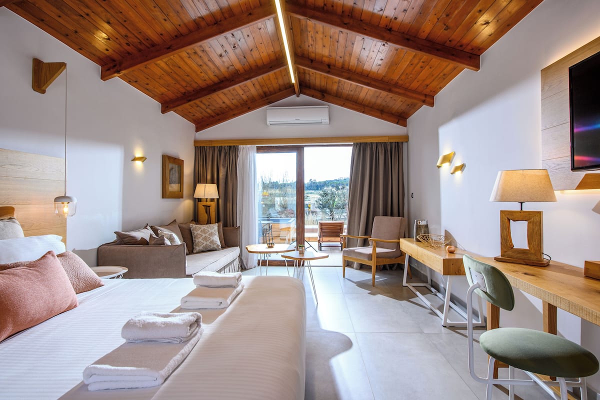 A hotel room with a wooden ceiling, a bed with white linens, a sofa, a desk with a chair, and a view of the outdoors through a large window.
