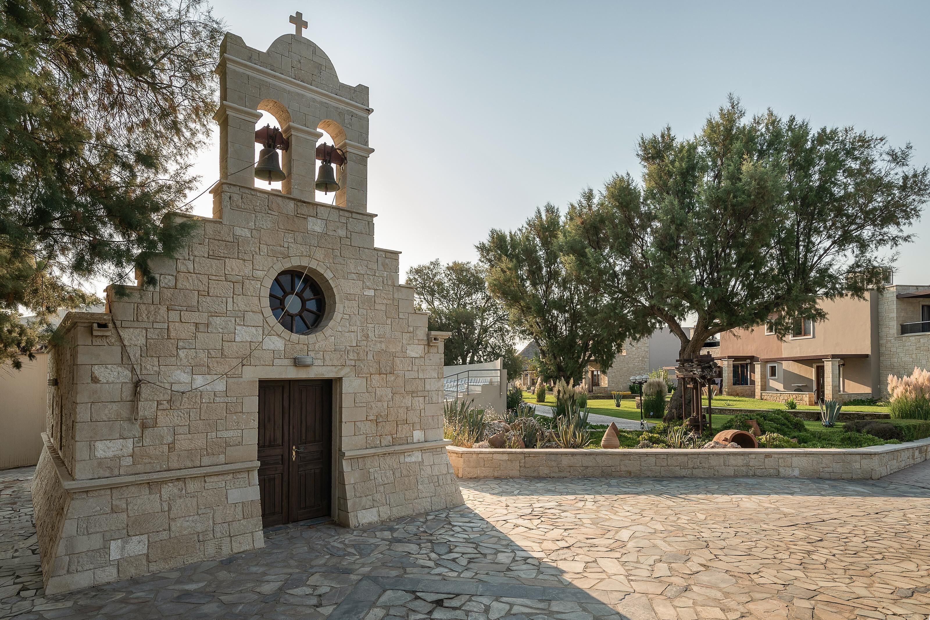 Una iglesia de piedra con un campanario, un reloj y un patio empedrado con un árbol grande.